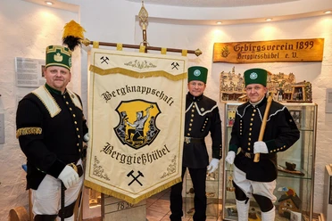 Three men in traditional miners' uniforms stand in front of a banner of the "Berggießhübel Miners' Association" in an exhibition room.