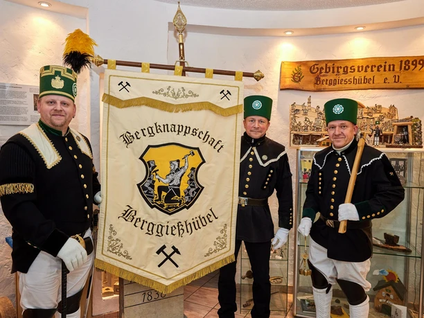 Drie mannen in traditionele mijnwerkersuniformen staan voor een spandoek van de "Berggießhübel Mijnwerkersvereniging" in een tentoonstellingsruimte.