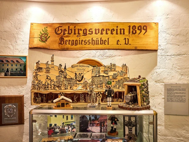 Vitrinekast met mijnbouwsouvenirs, waaronder een houten bord van de mijnbouwvereniging uit 1899, een mijnwerkersfiguur en historische foto's, verlicht in warm licht.