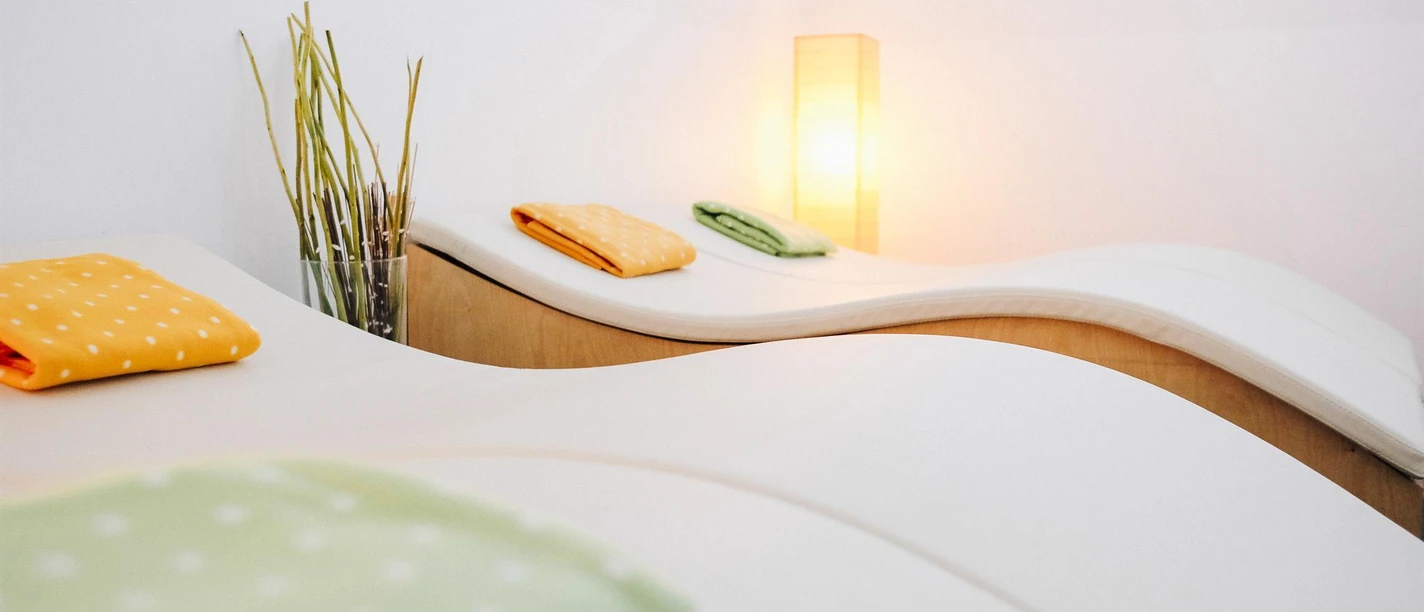 Heller Therapieraum mit Liegen, gefalteten Decken, Gräsern und warm leuchtender Lampe