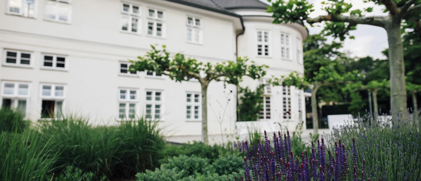 Weiße Salinenklinik mit dunklem Dach hinter blühendem Garten und Bäumen