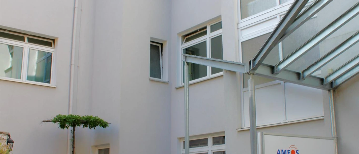 Eingang des AMEOS Klinikums mit weißer Fassade, Hinweisschild und blühender Bepflanzung