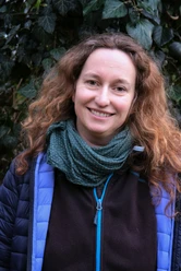 Hiking guide Daphna Zieschang A smiling woman with curly hair wears a blue jacket and a green scarf; green leaves can be seen in the background.