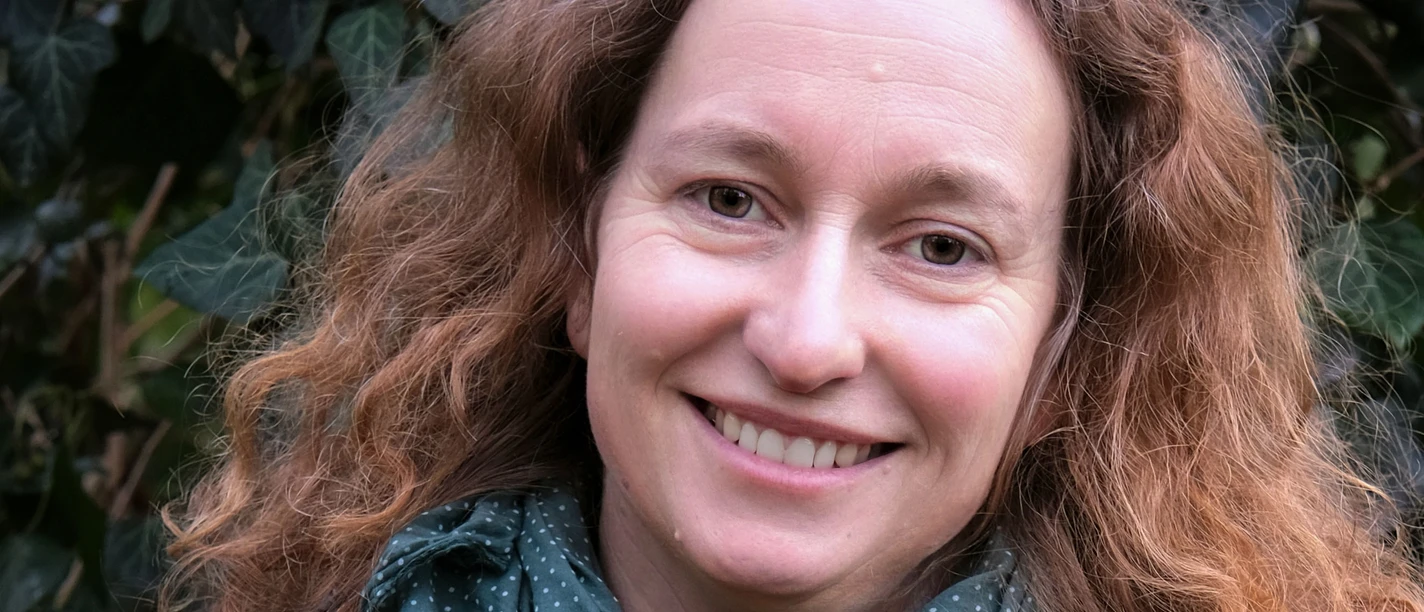 Wanderführerin Daphna Zieschang Eine lächelnde Frau mit lockigem Haar trägt eine blaue Jacke und einen grünen Schal; im Hintergrund sind grüne Blätter zu sehen.