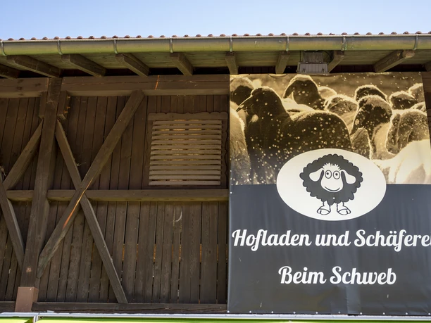 Hofladen beim Schweb Inser Hoamat Partner Hofladenplakat mit Schafsymbol, dunkelholzige Scheune im Hintergrund.