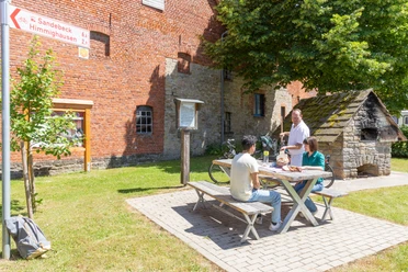 Nieheim-Oeynhausen-Backhaus-Teutoburger-Wald-Tourismus-A-Röser-100.jpg Drei Personen sitzen an einem Picknicktisch vor einem historischen Backstein- und Bruchsteingebäude.