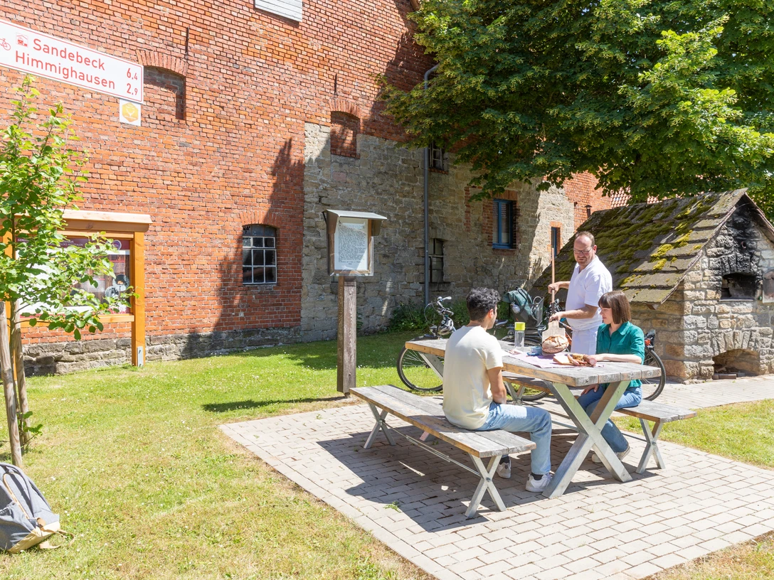 Nieheim-Oeynhausen-Backhaus-Teutoburger-Wald-Tourismus-A-Röser-100.jpg Drei Personen sitzen an einem Picknicktisch vor einem historischen Backstein- und Bruchsteingebäude.