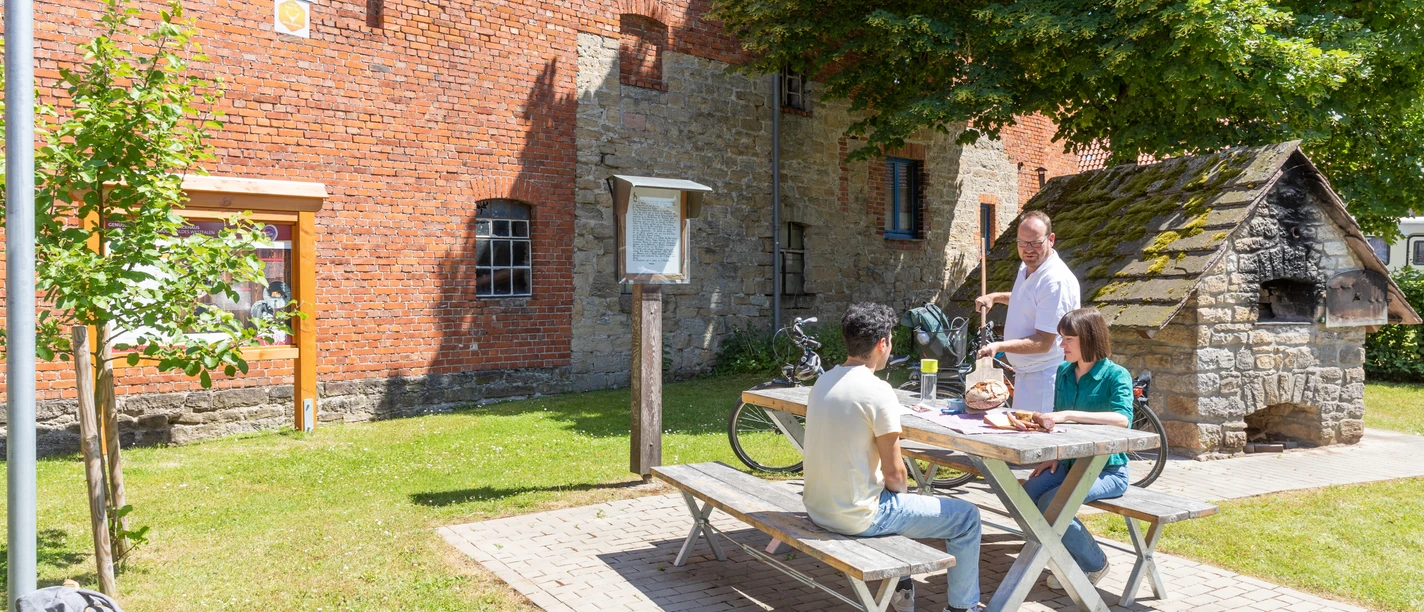 Nieheim-Oeynhausen-Backhaus-Teutoburger-Wald-Tourismus-A-Röser-100.jpg Drei Personen sitzen an einem Picknicktisch vor einem historischen Backstein- und Bruchsteingebäude.