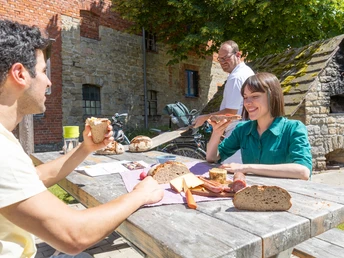 Drei Personen genießen ein Picknick an einem Holztisch vor einem rustikalen Gebäude bei Sonnenschein.