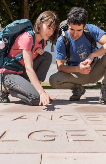 Zwei Wanderer betrachten Schriftzüge auf einer Steinplatte vor einem historischen Klostergebäude.