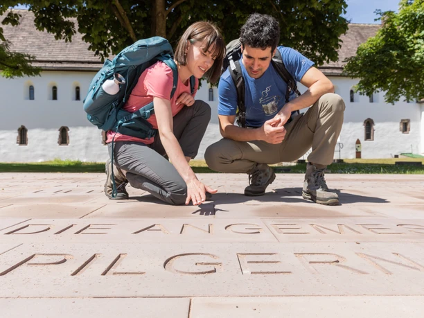 Höxter-Brenkhausen-Kloster-Teutoburger-Wald-Tourismus-A-Röser-042.jpg Zwei Wanderer betrachten Schriftzüge auf einer Steinplatte vor einem historischen Klostergebäude.