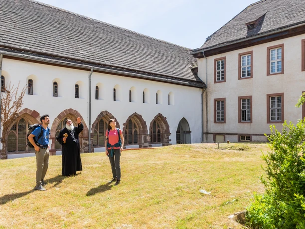 Klausenhof mit Besuchergruppe und Mönch im Gespräch, umgeben von historischer Architektur und grünem Rasen.