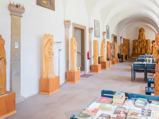 Blick in einen Flur mit hölzernen Skulpturen an den Wänden und einem Tisch voller Bücher im Vordergrund.