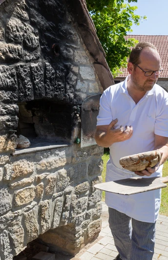 Ein traditioneller Bäcker zeigt einer Gruppe das Brotbacken in einem alten Steinofen im Freien.