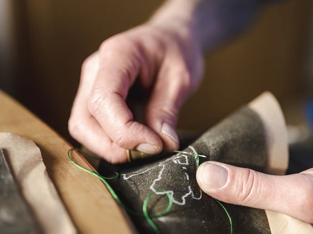 Lederhosen Michael Krippel Eine Hand näht mit grünem Faden ein Muster in dunkles Leder.