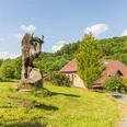 Steinbüffel-Skulptur auf grüner Wiese, Holzhütte und bewaldeter Hügel im Hintergrund.