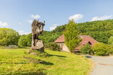 Warburg-Hammerhof-Teutoburger-Wald-Tourismus-A-Röser-051-CC-BY-SA.jpg Steinbüffel-Skulptur auf grüner Wiese, Holzhütte und bewaldeter Hügel im Hintergrund.