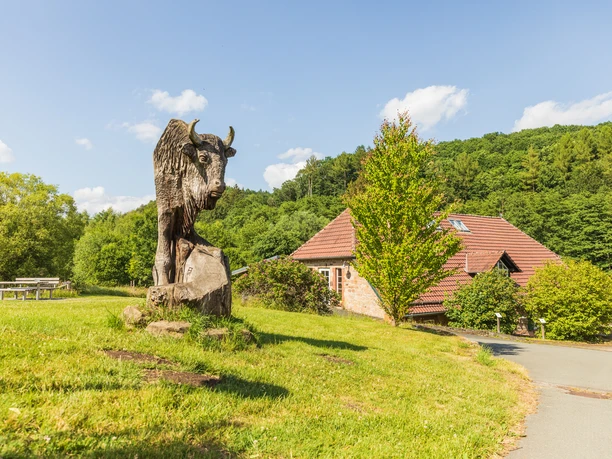 Warburg-Hammerhof-Teutoburger-Wald-Tourismus-A-Röser-051-CC-BY-SA.jpg Steinbüffel-Skulptur auf grüner Wiese, Holzhütte und bewaldeter Hügel im Hintergrund.