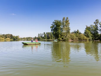 Ein grünes Ruderboot mit zwei Personen auf einem ruhigen See, umgeben von üppigen Bäumen.