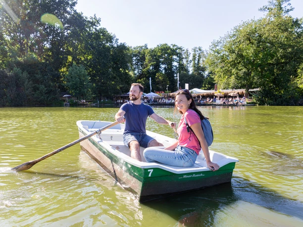 Spenge-Hücker-Moor-Teutoburger-Wald-Tourismus-A-Röser-157.jpg Ein Paar rudert bei Sonnenschein in einem grünen Boot über einen idyllischen See, umgeben von Bäumen.