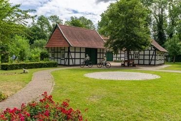 Hille-Kurpark-Rothenuffeln-Teutoburger-Wald-Tourismus-Patrick-Gawandtka-045.jpg Fachwerkhäuser in grüner Parklandschaft mit Radfahrern auf einem sonnenbeschienenen Weg.