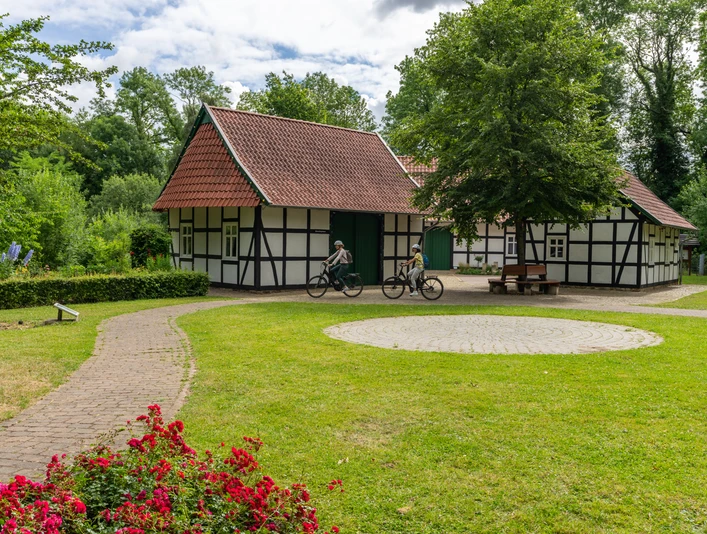 Hille-Kurpark-Rothenuffeln-Teutoburger-Wald-Tourismus-Patrick-Gawandtka-045.jpg Fachwerkhäuser in grüner Parklandschaft mit Radfahrern auf einem sonnenbeschienenen Weg.
