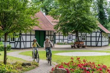 Hille-Kurpark-Rothenuffeln-Teutoburger-Wald-Tourismus-Patrick-Gawandtka-049.jpg Zwei Radfahrer auf einem Gartenweg vor einem Fachwerkhaus mit roten Dachziegeln und umgeben von Bäumen.