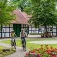 Hille-Kurpark-Rothenuffeln-Teutoburger-Wald-Tourismus-Patrick-Gawandtka-049.jpg Zwei Radfahrer auf einem Gartenweg vor einem Fachwerkhaus mit roten Dachziegeln und umgeben von Bäumen.