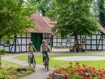 Hille-Kurpark-Rothenuffeln-Teutoburger-Wald-Tourismus-Patrick-Gawandtka-049.jpg Zwei Radfahrer auf einem Gartenweg vor einem Fachwerkhaus mit roten Dachziegeln und umgeben von Bäumen.
