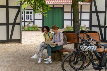 Hille-Kurpark-Rothenuffeln-Teutoburger-Wald-Tourismus-Patrick-Gawandtka-050.jpg Zwei Frauen sitzen auf einer Bank vor einem Fachwerkhaus, umgeben von Bäumen und Fahrrädern.