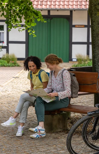 Hille-Kurpark-Rothenuffeln-Teutoburger-Wald-Tourismus-Patrick-Gawandtka-050.jpg Zwei Frauen sitzen auf einer Bank vor einem Fachwerkhaus, umgeben von Bäumen und Fahrrädern.