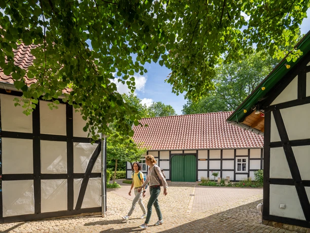 Hille-Kurpark-Rothenuffeln-Teutoburger-Wald-Tourismus-Patrick-Gawandtka-055.jpg Zwei Menschen spazieren durch einen gepflasterten Hof, umgeben von traditionellen Fachwerkhäusern.