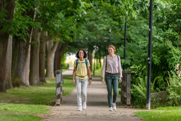 Hille-Kurpark-Rothenuffeln-Teutoburger-Wald-Tourismus-Patrick-Gawandtka-069.jpg Zwei Frauen spazieren entspannt auf einem von Bäumen gesäumten Weg im Park inmitten grüner Natur.