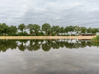Ein ruhiger See mit Wolkenhimmel und spiegelnder Wasseroberfläche, umgeben von Bäumen und Wiese.