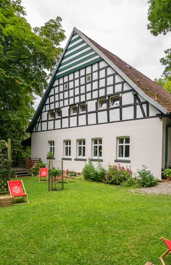 Huellhorst-CafeWaldkristall-Teutoburger-Wald-Tourismus-Patrick-Gawandtka-086-CC-BY-SA.jpg Fachwerkhaus im Grünen mit Liegestühlen im Vordergrund, umgeben von Bäumen und einer Terrasse.