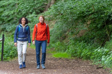 Huellhorst-Wandern-Teutoburger-Wald-Tourismus-Patrick-Gawandtka-074.jpg Zwei Frauen wandern lächelnd auf einem Waldweg, umgeben von üppigem grünen Laub.