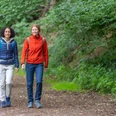Huellhorst-Wandern-Teutoburger-Wald-Tourismus-Patrick-Gawandtka-074.jpg Zwei Frauen wandern lächelnd auf einem Waldweg, umgeben von üppigem grünen Laub.