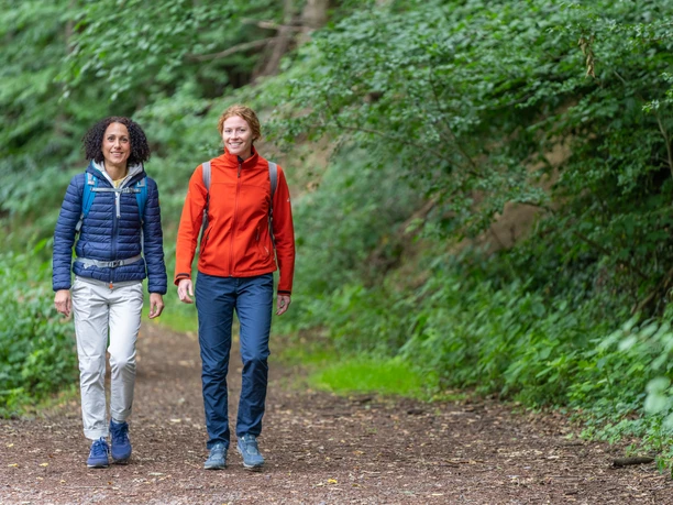 Huellhorst-Wandern-Teutoburger-Wald-Tourismus-Patrick-Gawandtka-074.jpg Zwei Frauen wandern lächelnd auf einem Waldweg, umgeben von üppigem grünen Laub.