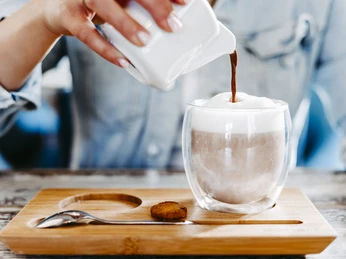Heimathafen Hand gießt heiße Schokolade in ein Glas mit Milchschaum, serviert auf einem Holztablett mit Keks.Hand pours hot chocolate into a glass with milk foam, served on a wooden tray with a cookie.Hånden hælder varm chokolade i et glas med mælkeskum, serveret på en træbakke med en kiks.Handgeschonken warme chocolademelk in een glas met melkschuim, geserveerd op een houten dienblad met een koekje.