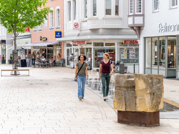 Zwei Frauen spazieren durch eine Fußgängerzone mit Geschäften und einem Café, bei sonnigem Wetter.