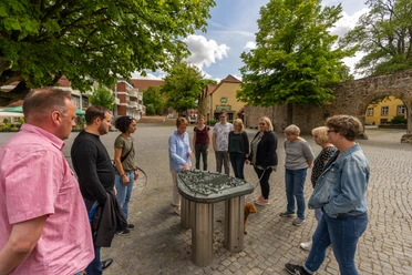 Menschen stehen um ein taktiles Modell einer historischen Stadt versammelt, umgeben von Bäumen und Gebäuden.