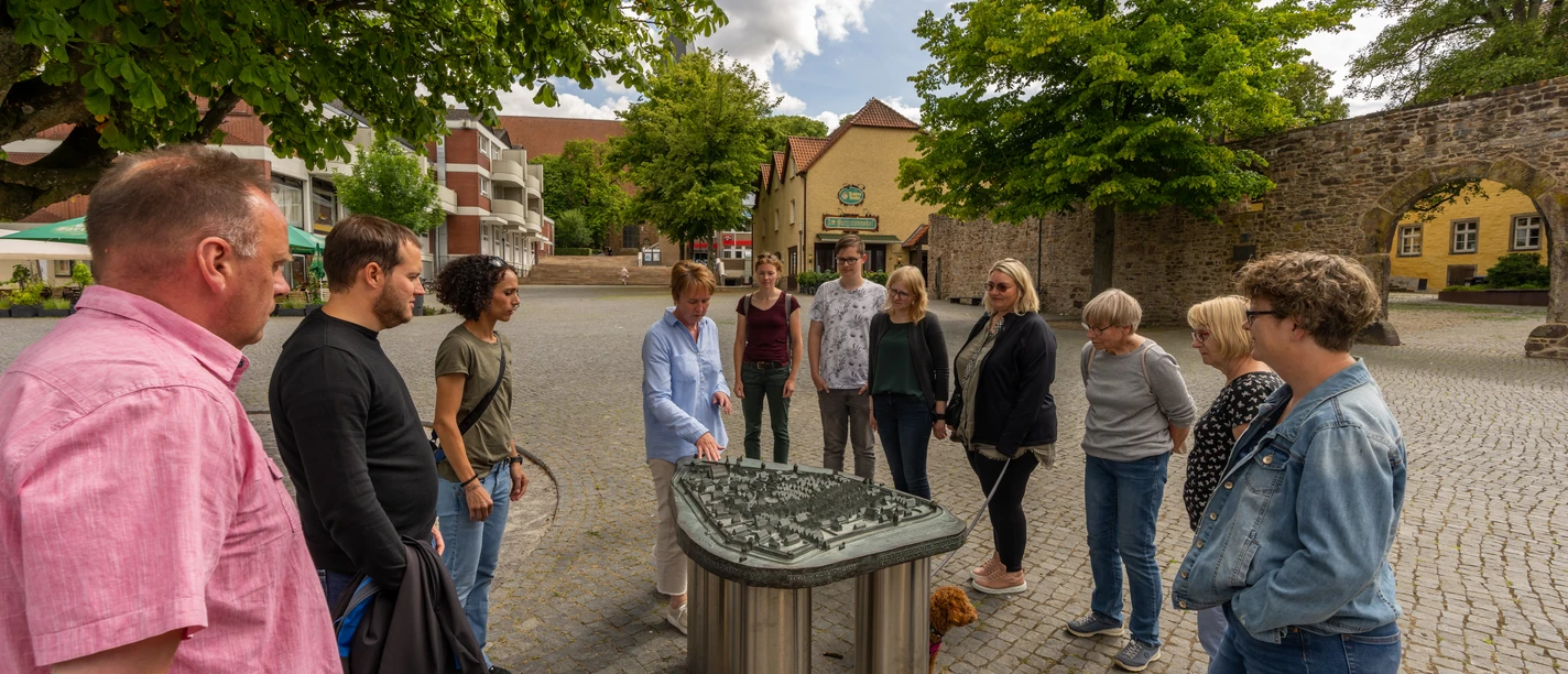 Menschen stehen um ein taktiles Modell einer historischen Stadt versammelt, umgeben von Bäumen und Gebäuden.