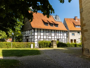 Fachwerkhaus an einem sonnigen Tag, umgeben von gepflastertem Weg und einigen Bäumen im Vordergrund.