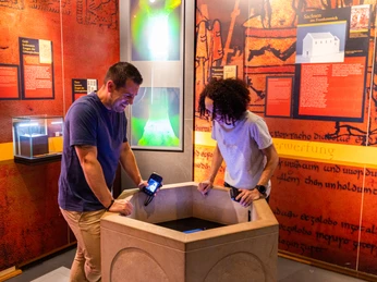 Zwei Personen betrachten im Museum einen historischen Taufstein, umgeben von Informationstafeln.
