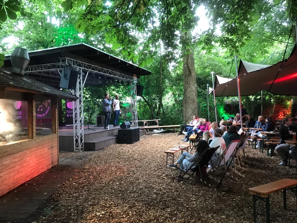 Büdchen am Westerberg.jpg Eine Open-Air-Bühne mit Zuschauern, umgeben von üppigen Bäumen in einem stimmungsvollen Setting.