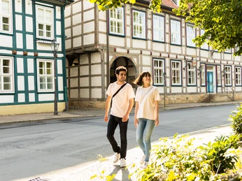 Ein Paar geht Hand in Hand durch eine idyllische Fachwerkstraße mit grünen Bäumen im Sonnenschein.