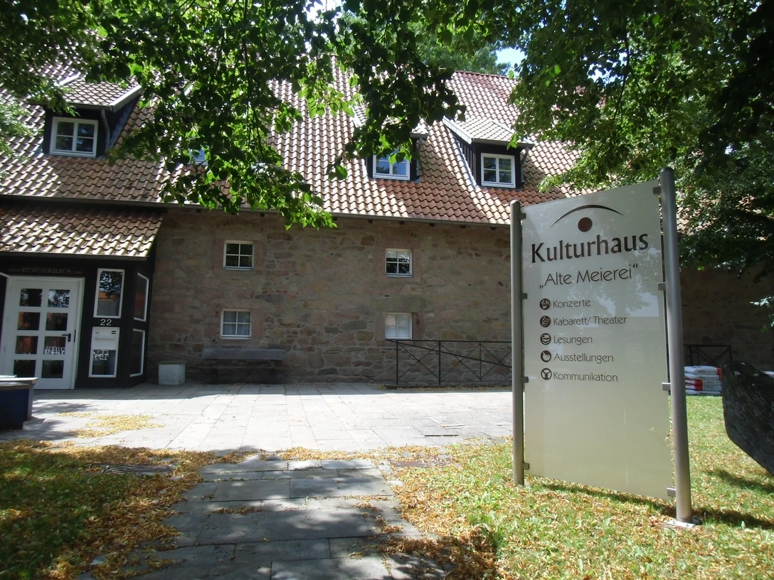Kulturhaus "Alte Meierei" Kulturhaus "Alte Meierei" mit Sandsteinfassade, Schildern für Veranstaltungen und schattigem Eingangsbereich.