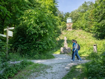 Ein Paar spaziert auf einem Waldweg vorbei an einem neoklassischen Denkmal im sommerlichen Grün.