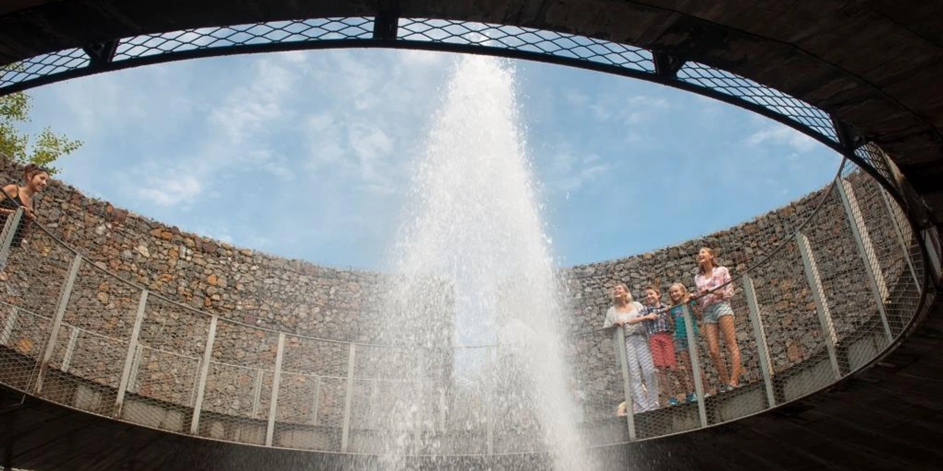 Wasserkrater auf der Aqua Magica Auf dem Bild ist der Wasserkrater in Aktion zu sehen, wobei das Wasser hochspritzt. Eine Familie steht neugierig am Geländer und beobachtet das beeindruckende Schauspiel.