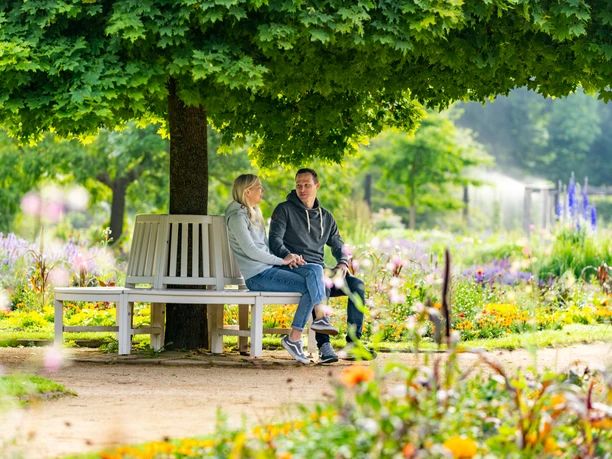 Guetersloh-BotanischerGarten-Teutoburger-Wald-Tourismus-Patrick-Gawandtka-016.jpg Zwei Personen sitzen entspannt auf einer Bank unter einem großen, schattenspendenden Baum in einem blühenden Garten.